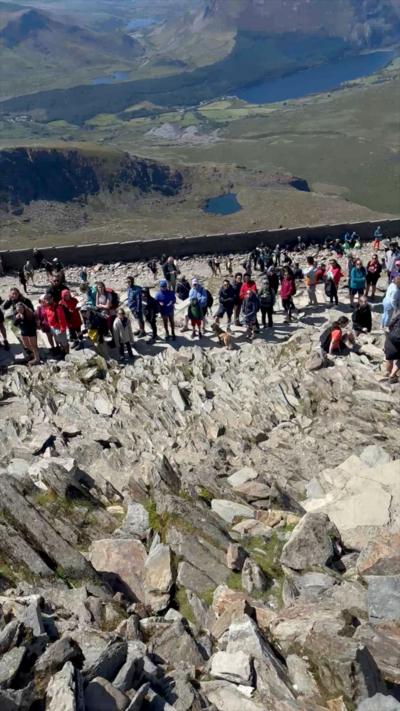 People queuing at the summit of Yr Wyddfa. They are below where the camera is and you can see rocks in the foreground and a lake in a valley behind