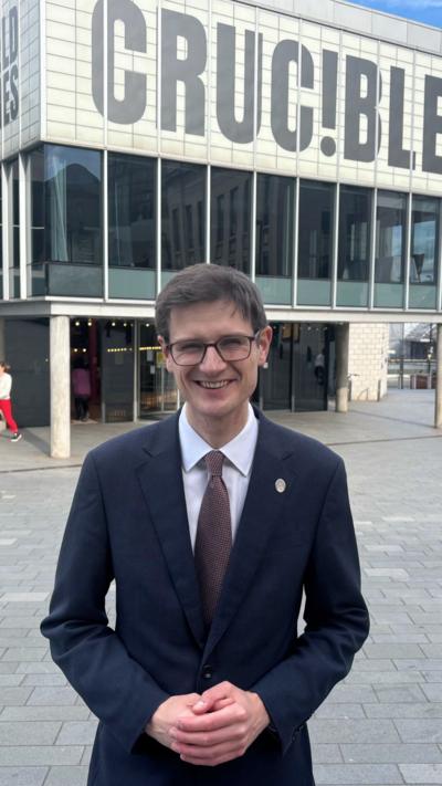 Tom Hunt, wearing a dark suit, white shirt and tie, smiles at the camera as he stands with his hands clasped in front of him outside Sheffield's Crucible Theatre.