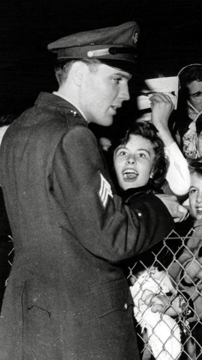 A black and white photo of Elvis on one side of a chain-link fence, with excited fans on the other.