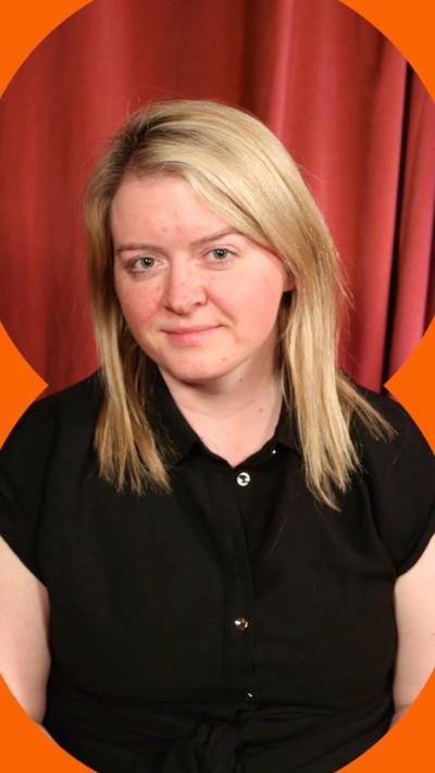 A woman with blonde shoulder-length hair and a black shirt sits on a chair, in front of a red curtain.
