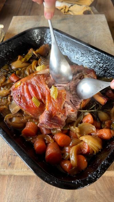A lamb joint surrounded by vegetables in black baking tin