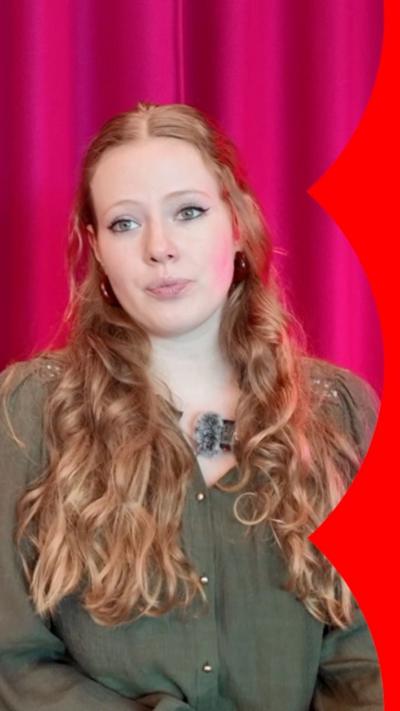 A woman with long red wavy hair wears a khaki coloured top and sits against a pink curtain