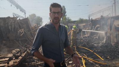 Dan Johnson wears a dark shirt and glasses, standing amongst rubble with smoke in the air and firefighers using a hose in the background