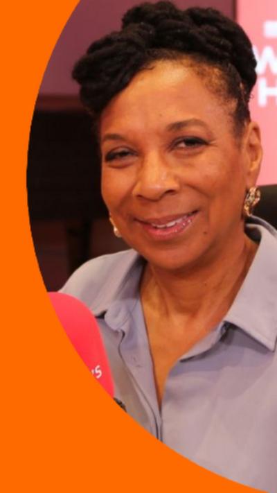 Kimberle Crenshaw has dark hair and is wearing a light purple shirt. She is sat infant of a red radio microphone.