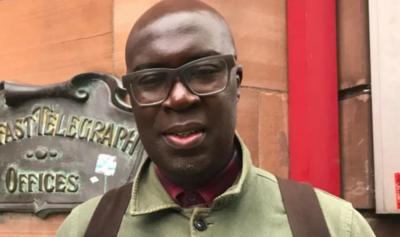 Kwame Daniels in front of the Telegraph building in Belfast. He is wearing thick-rimmed glasses, a green jacket, a red shirt and a ruck sack.