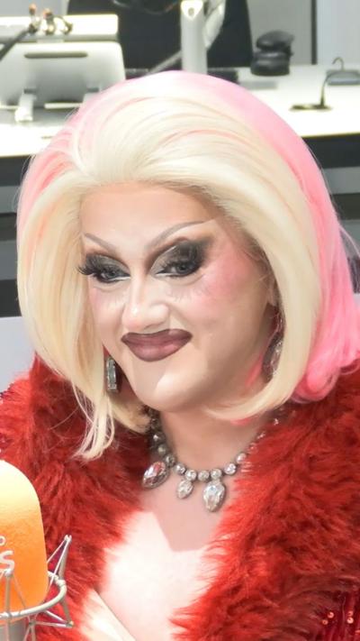 Catrin Feelings, who is in heavy drag make-up and a blonde wig which is pink at the back, sat in a red feathery coat and silver necklace