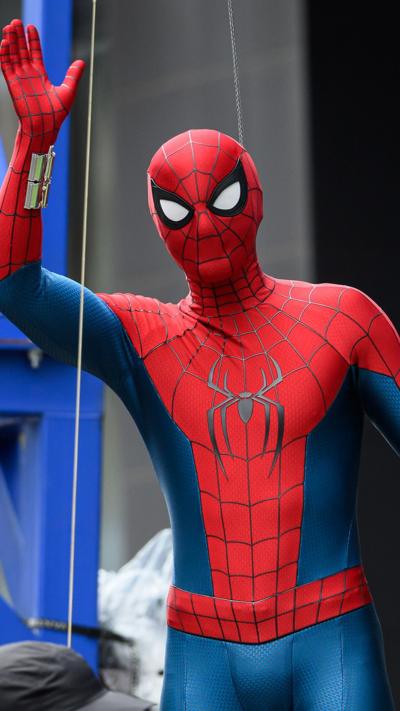 Tom Holland wearing Spider-Man bodysuit and mask waves while strapped to stunt wires and crane