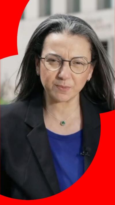 Shaimaa Khalil has long dark hair and is wearing glasses, a black blazer and a blue top.