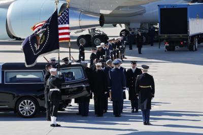 People in military dress stand in line next to a hearse and aeroplane with two large flags.