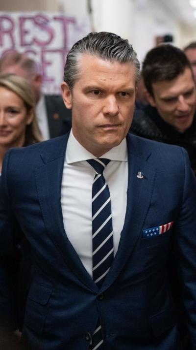 Pete Hegseth walks wearing a blue suit and striped tie