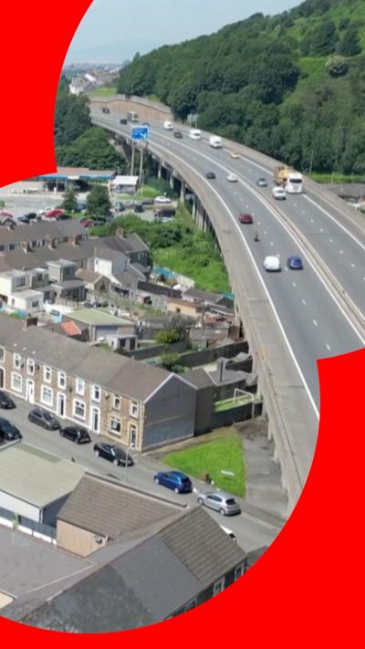 The M4 motorway can be seen on the right hand side of the image, on the left hand side is a tightly arranged group of houses. The motorway is raised above the land that the houses stand on, it has pillars that keep it elevated that overhangs some of the residential land, some of pillars that keep it elevated have been erected in the gardens of these houses. There are also more houses that stretch all the way to the horizon, as well as a massive cluster of trees on a hill. 
