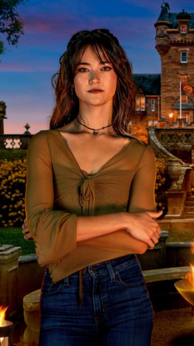 Woman with long dark hair which is pinned up behind her head with a long fringe at the front brushed away to each side, she is smiling and wearing long red tassel style earrings and two delicate necklaces, one of which has a green gemstone. The off the shoulder outfit is in navy and dark green tartan styled like a bow on the right side. She is standing in front of a blue bookcase which has books on the shelves.