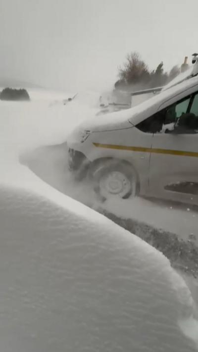 The front of a parked van is surrounded by a high wall of snow, which is still falling.