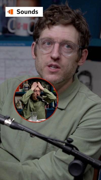 Comedian Elis James sits in a recording studio, he wears a green shirt and translucent glasses