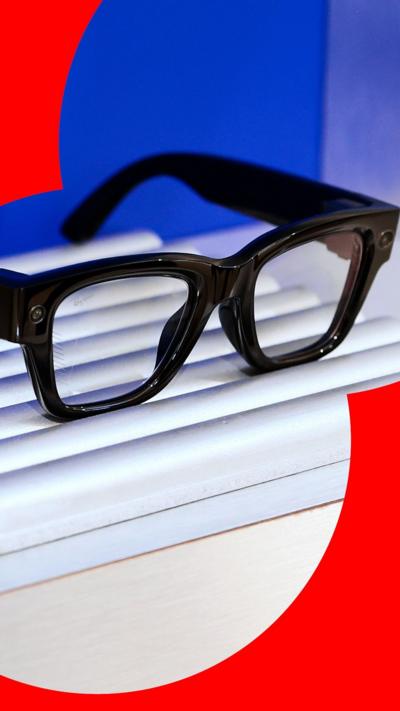 A pair of black-rimmed smart glasses on a display stand. They have two cameras, one in the corner of each frame.
