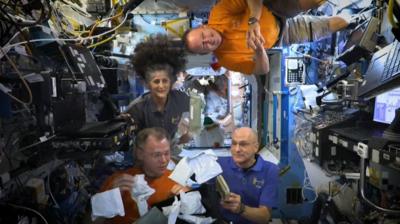 Four astronauts float next to screens and wires on the ISS while unboxing white packages