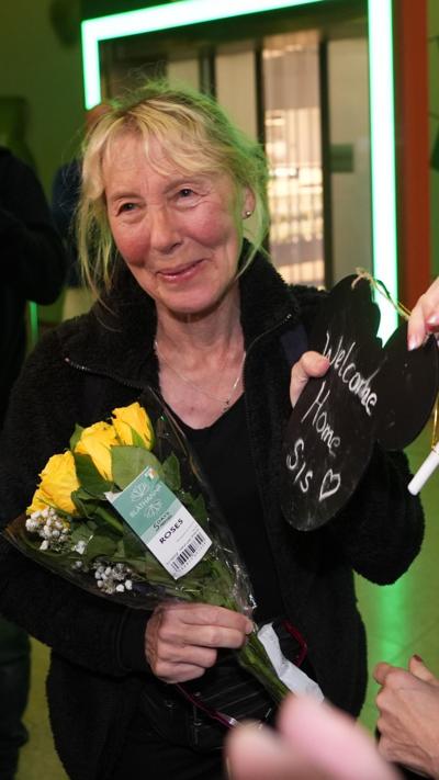 a woman smiling while holding a "welcome home sis" sign along with yellow roses