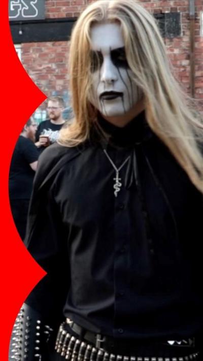 Envus is stood in front of a crowd outdoors. He is wearing all black, has long brown hair and his face painted white with black accents.
