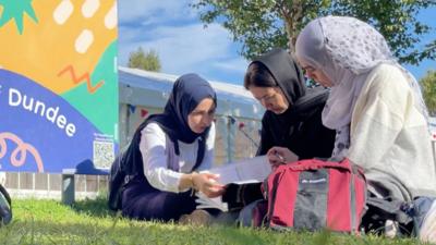 Three women sitting on grass looking at coursework together. They are near a sign that says Dundee.