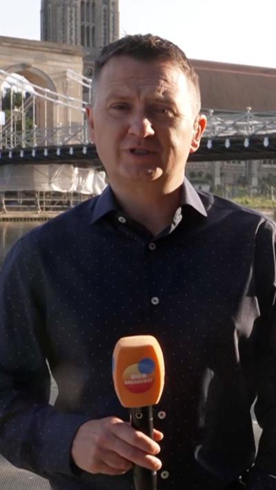 Matt Taylor in a black shirt, holding a microphone and stood by a riverside