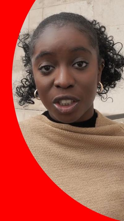 A woman with curly black hair is wear a camel-coloured wrap. She is speaking into the camera and holding a button mic.