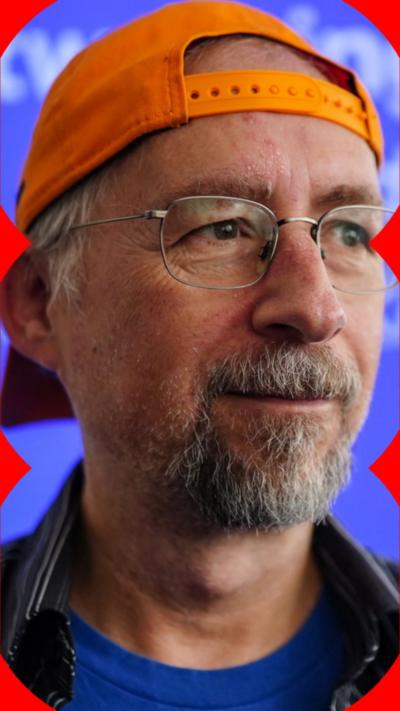 Adam Back is wearing an orange cap in reverse and has silver-framed glasses.