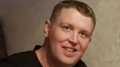 Robert Kincaid has short blond/brown hair. He is smiling and is wearing a black top. The back drop looks like wallpaper.