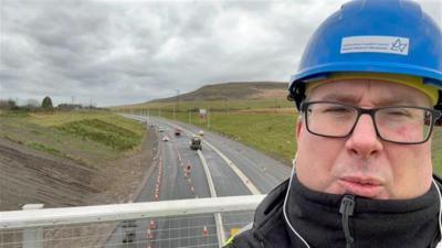 BBC Wales Journalist Pete Shuttleworth in front of the heads of the valleys road works. He is wearing a helmet. 