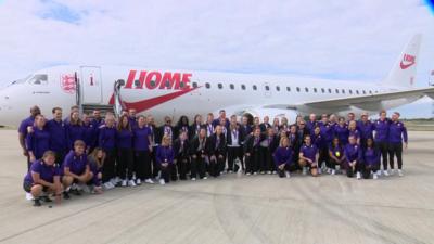 The Lioness team stood in front of a jet that has a Nike symbol and the word 'Home' on it