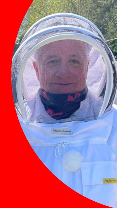 A man smiles at the camera. He is wearing a fully-enclosed beekeeping suit and hood, and a blue scarf with pink squiggles around his neck.