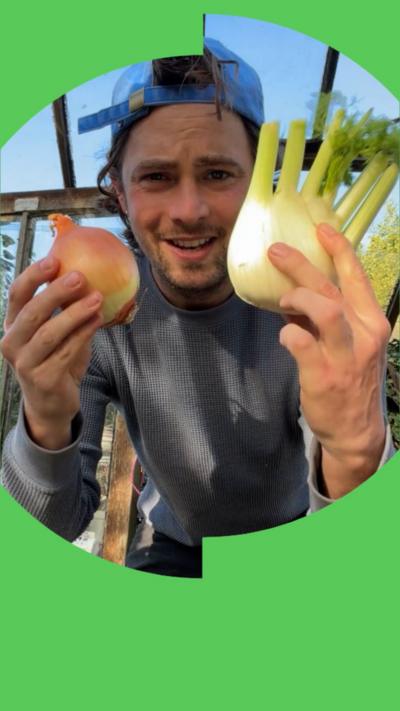Julius Roberts holds up an onion in his right hand and a head of fennel in his left. He is wearing a grey sweatshirt and a blue baseball cap on backwards.