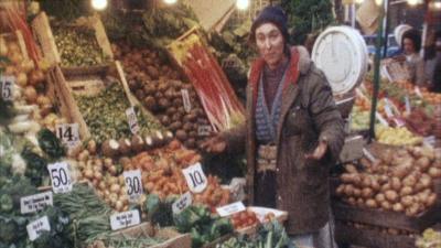 A stallholder wearing a brown coat and blue hat gestures next to her vegetable stall.