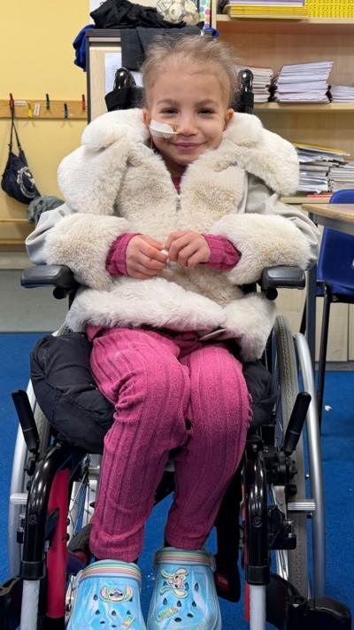 Skye, a young girl sitting in a wheelchair and smiling. She is wearing a fluffy jacket and pink leggings, with blue Crocs.