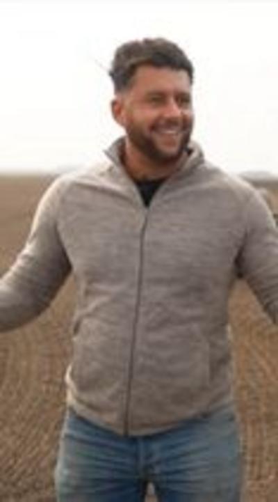 A man stands in a ploughed field smiling and looking off camera to the right. He has got dark hair and a beard, and is wearing a grey zip up top and blue jeans.