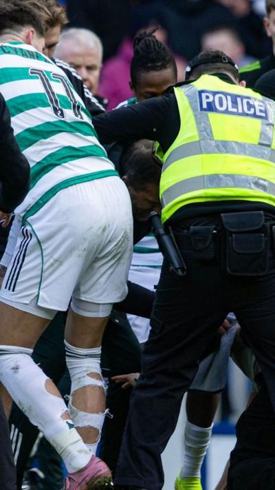 Fans invade pitch at Ibrox, Celtic player with torn socks and a police officer can be seen in the crowd
