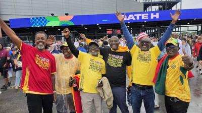 Six men stand in a line smiling and looking excited as they raise their arms in the air, wave at the camera and give a thumbs up. They are all wearing some combination of Ghana colours, predominantly yellow. Behind them, a crowd moves outside a stadium, with a blue video sign above an entrance which reads 'Gate H' in capital letters 