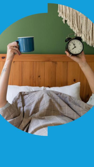 Two arms stick up from under a duvet. One is holding a clock, the other is holding a mug