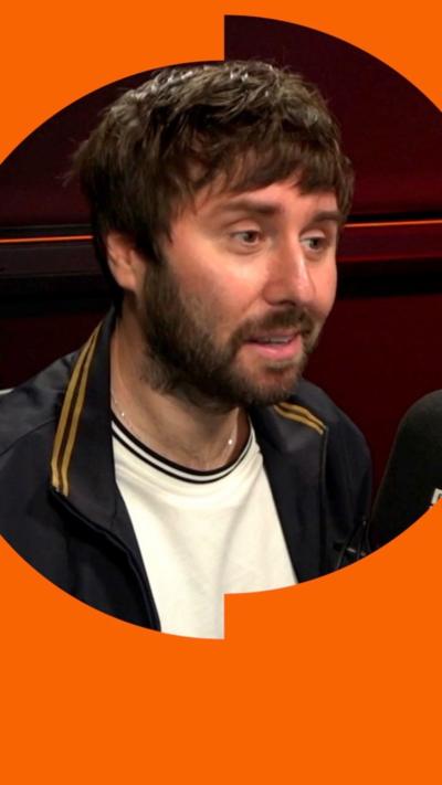 James Buckley, smirking, wearing white t-shirt and black jacket. He has a beard and dark hair.