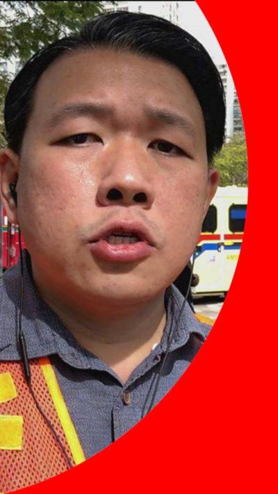 A man wearing an orange jacket over a shirt, with an ambulance and fire engine behind.