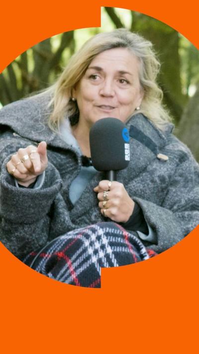A woman wrapped up in a coat and blanket, holding a mic, in a forest.