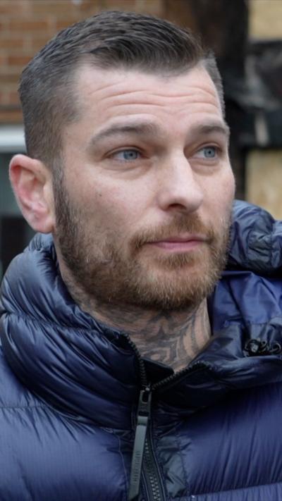 Dean Ray is wearing a blue puffer jacket which is hooded. He is looking off camera to the right. He has a short, brown haircut with a facial hair. He had a neck tattoo.