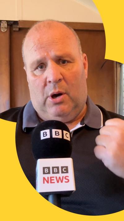 Alternative description
A close-up of content creator Big John wearing a black polo and holding a microphone with the words 'BBC News'