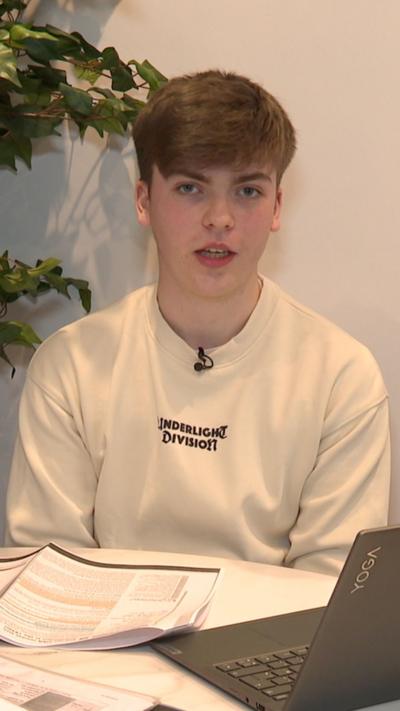 A young man with brown hair and a fringe in a cream jumper sat in front of a laptop with papers 