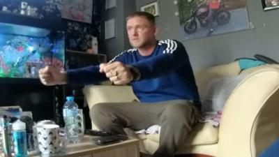 A man sat on a sofa with his arms in front of him. There is a photo behind him on the wall, a fish tank to his right and bottles and glasses on the table.