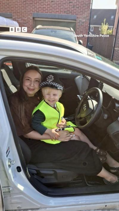 A woman sits a in car with a young child on her lap. He is wearing a hi-vis jacket and a police hat.