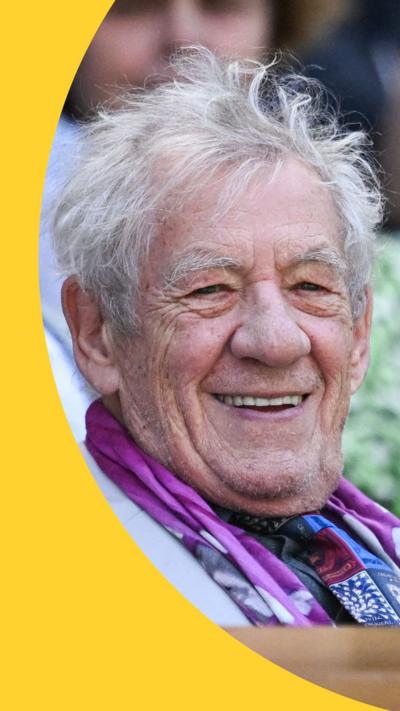 Ian McKellen smiles, sitting in the stands at Wimbledon Centre Court. He has short grey hair and wears a purple neck scarf