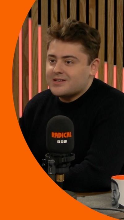 Joe Seddon has dark hair and is wearing a black long-sleeved top. He sits behind a mic