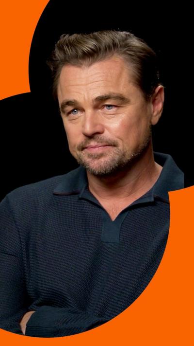 Actor Leonardo DiCaprio sat in front of a plain black background. He has his arms folded and a long sleeved blue shirt on