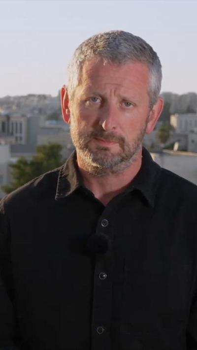 Jon Donnison stands in a black shirt with Jerusalem skyline behind him