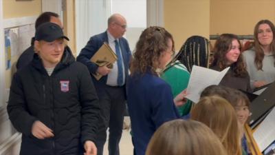 Ed Sheeran, wearing a baseball cap and a padded winter coat, smiles after walking into a room to hear a choir. The young members of the choir are smiling and singing.